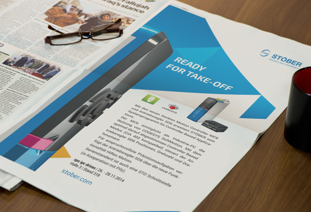 Publicité Stober sur une table avec journal, lunettes et tasse. Texte : "READY FOR TAKE-OFF".